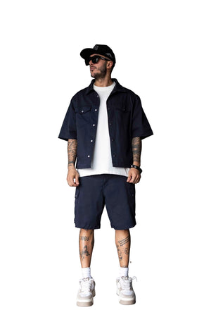 A man stands in a studio wearing a dark navy short-sleeve button-up shirt over a white t-shirt, matching navy shorts, white sneakers, and a black cap.