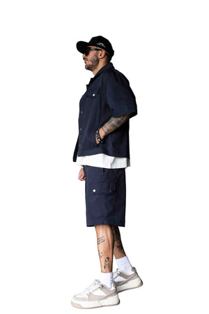 A man wearing a navy blue short-sleeve button-up shirt, matching cargo shorts, a white undershirt, white socks, and beige sneakers, standing in a modern gray room.