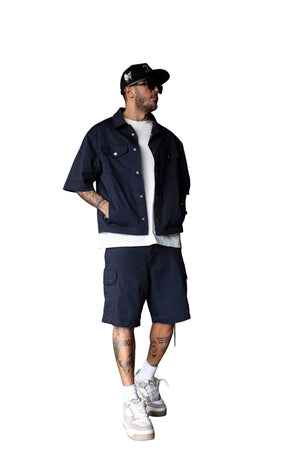 A man wearing a navy blue short-sleeve button-up shirt, matching navy cargo shorts, a white t-shirt, white sneakers, and a black baseball cap, standing in a modern industrial room with a concrete wall.