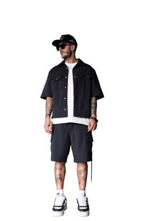 A man wearing a black short-sleeve button-up shirt, black cargo shorts, a white t-shirt, black and white sneakers, and a black baseball cap, standing in a gray concrete room.