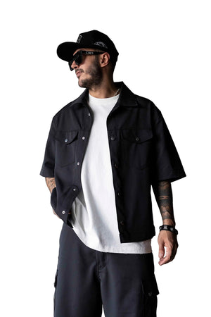 A man wearing a black short-sleeve button-up overshirt over a white t-shirt, paired with black cargo pants, a black cap, and sunglasses, standing against a textured gray wall.