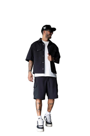 A man wearing a black short-sleeve button-up shirt, black cargo shorts, a white t-shirt, black and white sneakers, and a black baseball cap, standing in a gray industrial room.