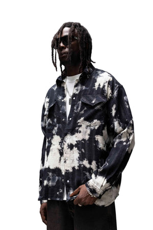 A man wearing a black and white tie-dye button-up shirt over a white t-shirt, styled with dark pants and sunglasses, standing in a modern concrete architectural setting.