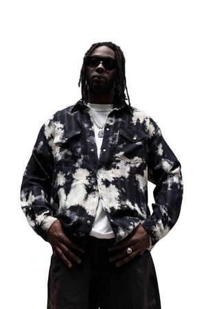 A man wearing a black and white tie-dye denim shirt jacket over a white t-shirt, styled with dark sunglasses, a silver chain necklace, and dark brown pants, standing against a concrete wall.
