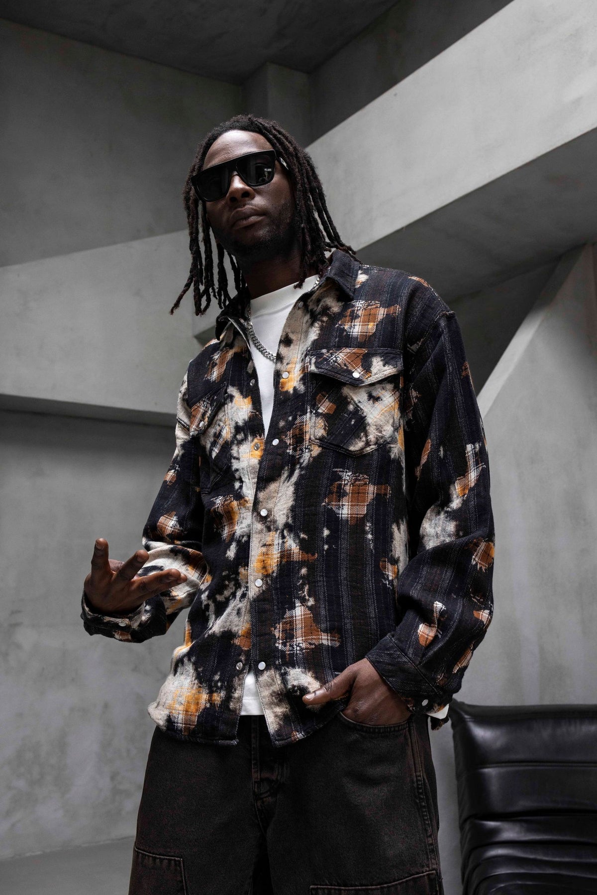 A man wearing a long-sleeve button-up shirt with a black, white, and orange abstract tie-dye pattern, styled with dark sunglasses, a white t-shirt, and dark cargo pants in a modern concrete setting.