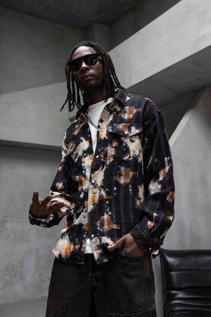 A man wearing a long-sleeve button-up shirt with a black, white, and orange abstract tie-dye pattern, styled with dark sunglasses, a white t-shirt, and dark cargo pants in a modern concrete setting.