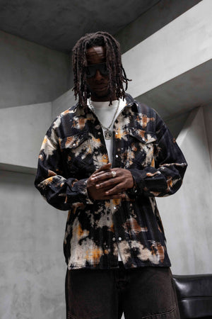 A man with long dreadlocks and sunglasses wearing a black button-up shirt with a brown and beige abstract tie-dye pattern, layered over a white t-shirt and dark pants, standing in a modern concrete room.