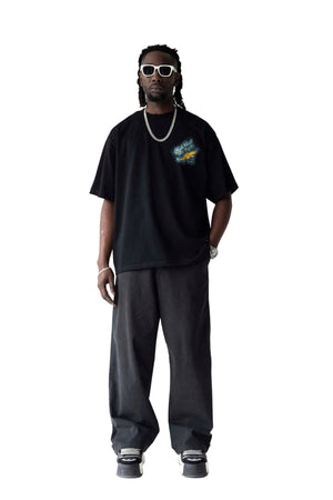 A man modeling a black oversized t-shirt with a colorful graphic on the left chest, paired with loose black sweatpants, white sunglasses, and a silver chain necklace, standing against a light gray studio background.