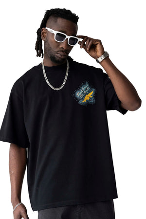 A man wearing an oversized black t-shirt with a blue and yellow graphic on the left chest, styled with white sunglasses, a silver chain necklace, and a silver watch, set against a plain light gray background.