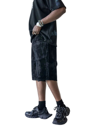A person shown from the waist down wearing black textured shorts, a black graphic t-shirt, black socks, and black chunky sneakers against a plain white studio background.