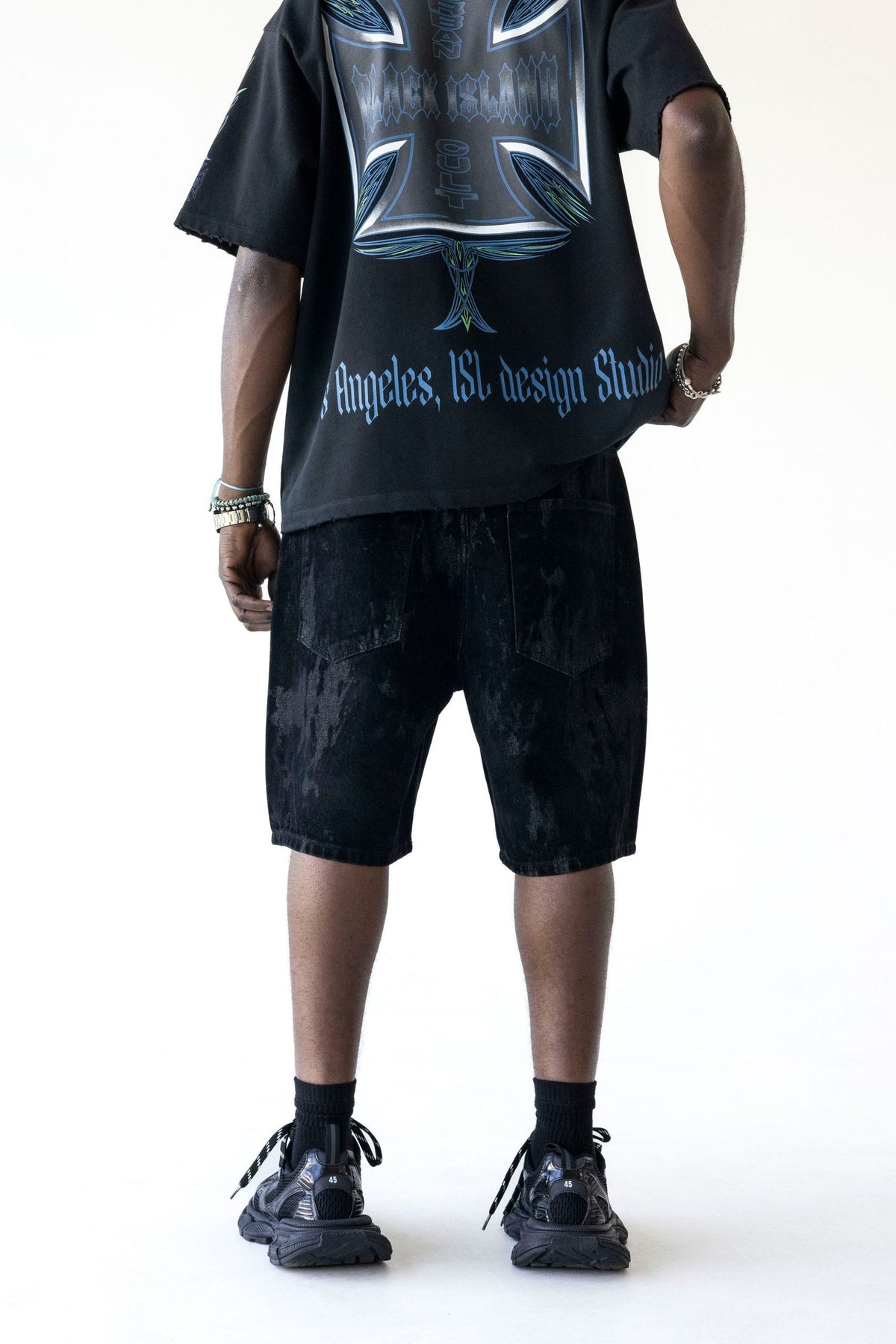 A model from the back wearing a black graphic t-shirt and black distressed denim shorts, paired with black socks and black sneakers, against a plain white background.