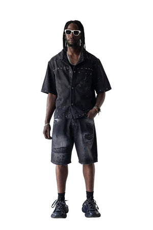 A man modeling a black short-sleeve button-up shirt and matching distressed black shorts, styled with white sunglasses, black socks, and black sneakers against a plain white studio background.