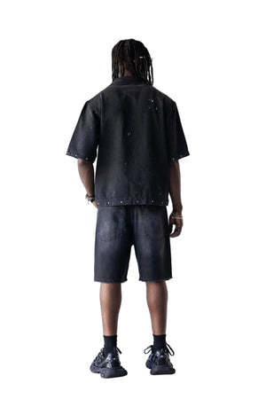 A model viewed from behind wearing a black short-sleeve button-up shirt and matching black denim shorts, styled with black socks and black sneakers against a plain white studio background.