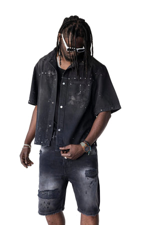 A man modeling a black distressed short-sleeve denim shirt and matching faded black denim shorts against a plain white studio background.