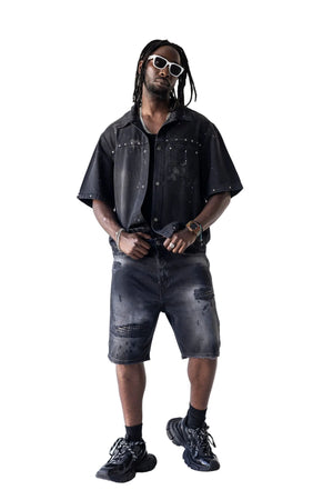 A man modeling a matching black distressed short-sleeve button-up shirt and distressed denim shorts, styled with black sneakers, black socks, and white-framed sunglasses against a white studio background.