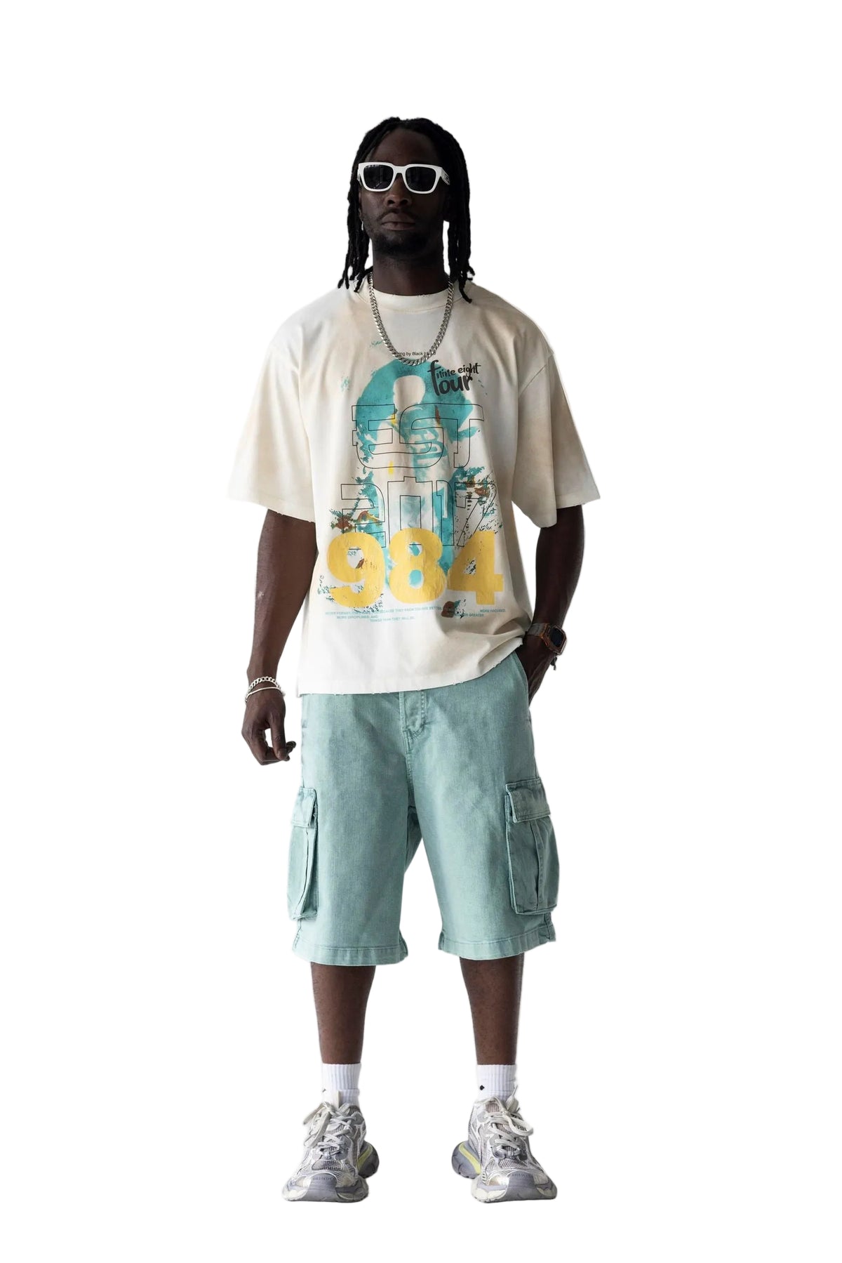 A man modeling a cream graphic t-shirt with a teal and yellow print, paired with light teal cargo shorts, white socks, and gray sneakers against a white studio background.