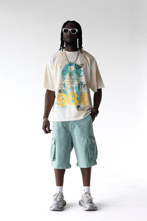 A man modeling a cream graphic t-shirt with a teal and yellow print, paired with light teal cargo shorts, white socks, and gray sneakers against a white studio background.