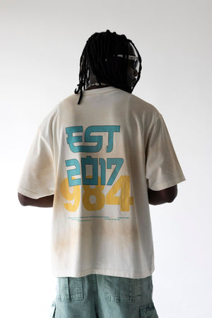 A person shown from the back wearing an oversized off-white graphic T-shirt with large teal and yellow text reading 'EST 2017 984' on the back, paired with light blue jeans against a plain light background.