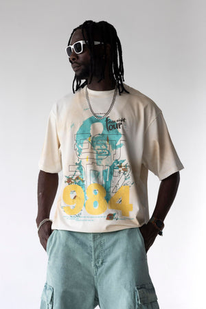 A man wearing a cream-colored graphic t-shirt with a blue and yellow '984' design, styled with light blue cargo shorts, white sunglasses, and a silver chain necklace against a light gray studio background.