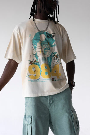 A person wearing an oversized cream graphic T-shirt with a large teal and yellow '984' design, paired with light blue cargo shorts against a plain light gray studio background.