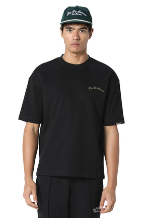 A man wearing a black crewneck t-shirt with a small gold embroidered logo on the left chest, paired with black pants and a dark green baseball cap. The background is a clean, light gray studio backdrop.