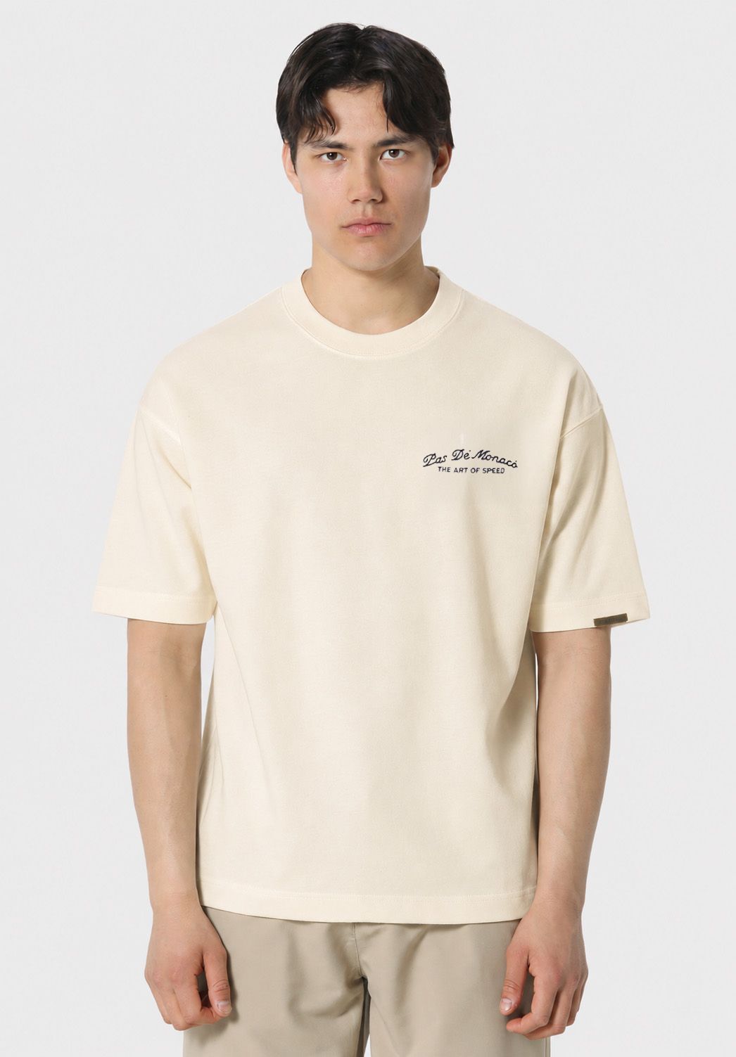 A man wearing a cream-colored short-sleeve t-shirt with a small black embroidered logo on the left chest, paired with beige pants, against a light gray background.