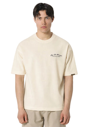 A man wearing a cream-colored short-sleeve t-shirt with a small black embroidered logo on the left chest, paired with beige pants, against a light gray background.