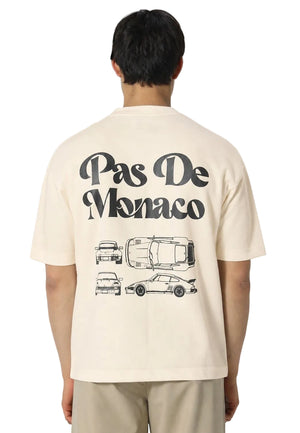 A man viewed from behind wearing a cream-colored t-shirt with a black graphic print that reads 'Pas De Monaco' above car illustrations, paired with beige trousers, against a light gray studio background.