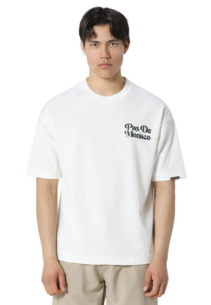 A man wearing a white short-sleeve crewneck t-shirt with a black 'Pas De Monaco' logo printed on the left chest, paired with beige trousers, against a light gray studio background.