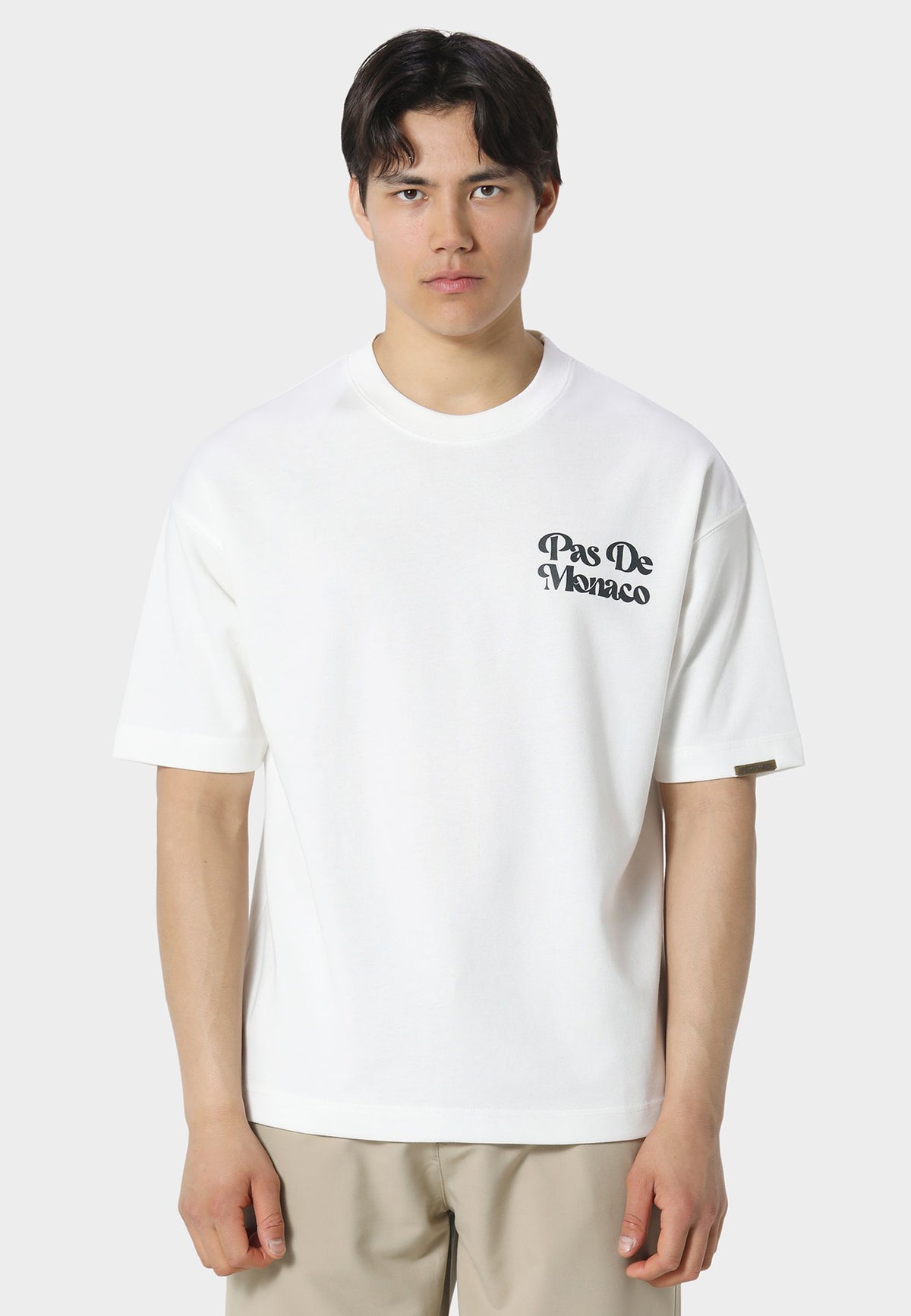 A man wearing a white short-sleeve crewneck t-shirt with a black 'Pas De Monaco' logo printed on the left chest, paired with beige trousers, against a light gray studio background.