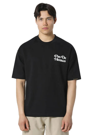 A man wearing a black short-sleeve crewneck t-shirt with 'Pas De Monaco' printed in white gothic font on the left chest, paired with beige trousers, against a light gray background.
