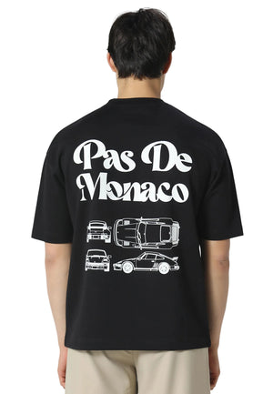 A man viewed from the back wearing a black t-shirt with a large white graphic print that reads 'Pas De Monaco' above four white car line drawings, paired with light beige pants against a light gray studio background.