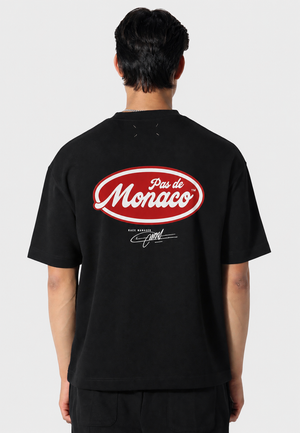 A man viewed from behind wearing a black t-shirt with a large red and white 'Pas de Monaco' logo printed on the back.