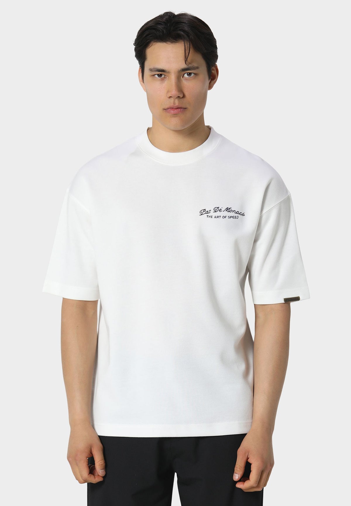 A man wearing a white short-sleeve crewneck t-shirt with a small black script logo on the left chest, paired with black pants, against a light gray studio background.