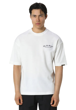 A man wearing a white short-sleeve crewneck t-shirt with a small black script logo on the left chest, paired with black pants, against a light gray studio background.
