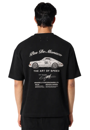 A man viewed from behind wearing a black t-shirt with a large white graphic print on the back. The graphic features a white sports car, the text 'Pas De Monaco', 'THE ART OF SPEED', and 'Gand' in a vintage style.