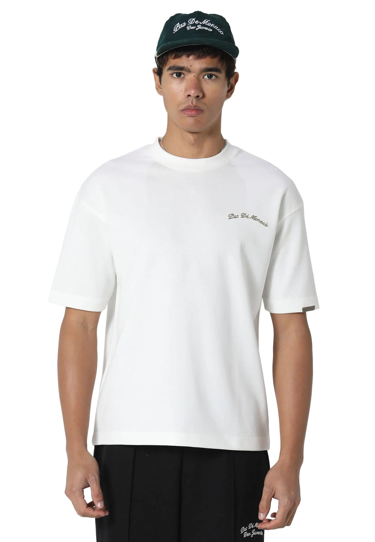 A man wearing a white short-sleeve crewneck t-shirt with a small cursive 'Balenciaga' logo on the left chest, paired with black pants and a dark green cap.