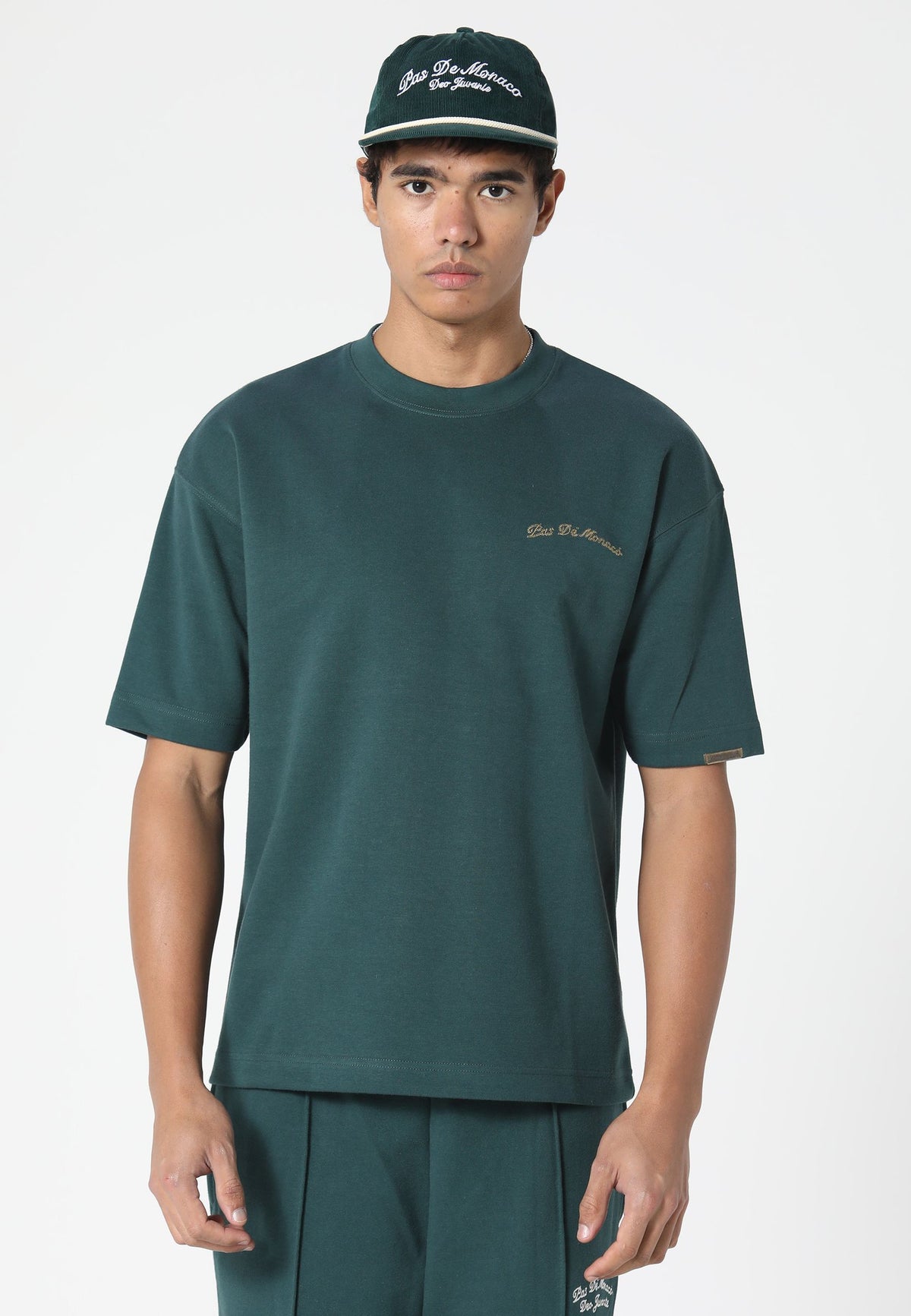 A male model wearing a dark green crewneck t-shirt with a small gold embroidered logo on the left chest, matching dark green pants, and a dark green baseball cap with white embroidery. The background is a clean, light gray studio backdrop.