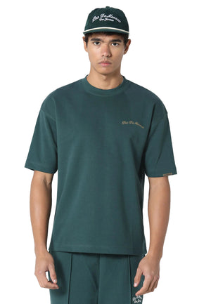 A male model wearing a dark green crewneck t-shirt with a small gold embroidered logo on the left chest, matching dark green pants, and a dark green baseball cap with white embroidery. The background is a clean, light gray studio backdrop.