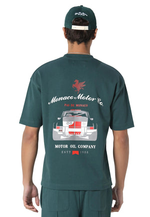 A man shown from the back wearing a dark green t-shirt with a large 'Monaco Motor Co.' racing graphic on the back, paired with matching green pants and a cap.
