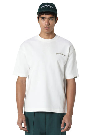 A man wearing a white short-sleeve crewneck t-shirt with a small brown embroidered logo on the left chest, paired with dark green pants and a dark green baseball cap.