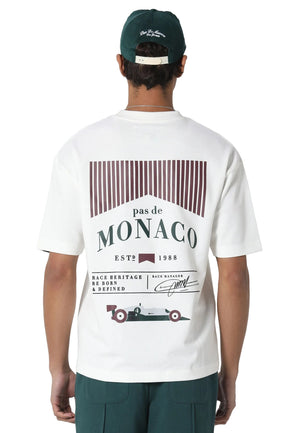 A man viewed from behind wearing a white t-shirt with a large 'pas de MONACO' graphic on the back, paired with a dark green cap and matching green pants, against a light gray studio background.