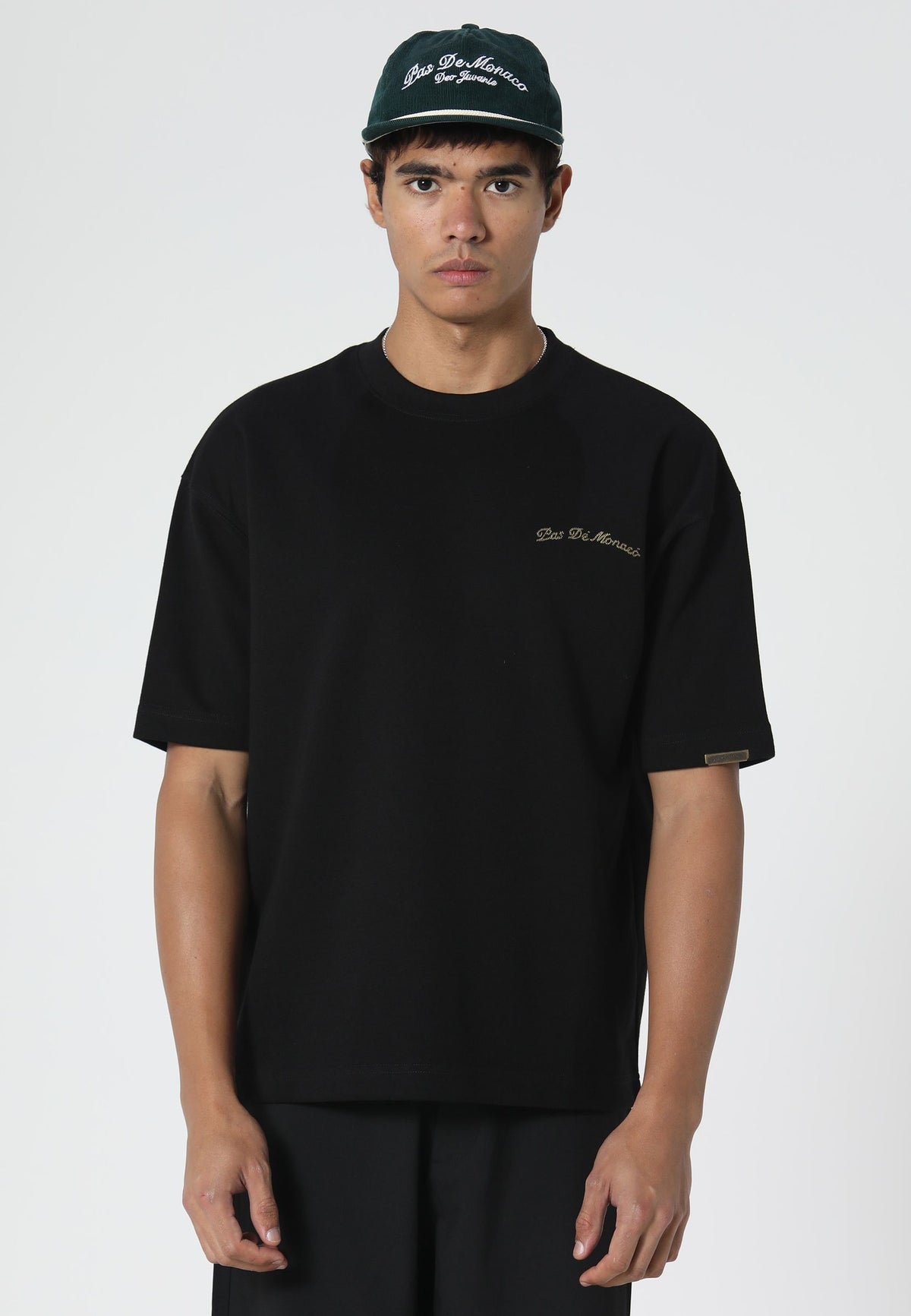 A man wearing a black short-sleeve t-shirt with a small gold embroidered logo on the left chest, paired with a black baseball cap. The background is a clean, light gray studio backdrop.