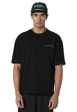 A man wearing a black short-sleeve t-shirt with a small gold embroidered logo on the left chest, paired with a black baseball cap. The background is a clean, light gray studio backdrop.