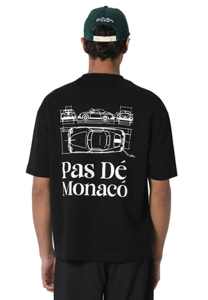 A man viewed from behind wearing a black t-shirt with a white graphic of sports cars and the text 'Pas Dé Monaco' on the back, paired with a dark green baseball cap and black pants, against a light gray studio background.
