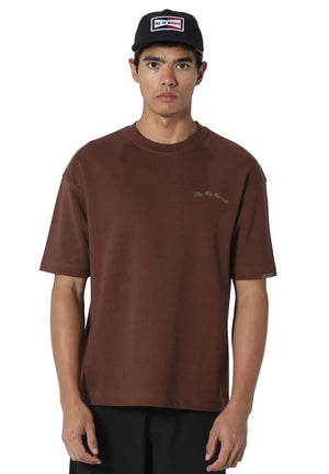 A man wearing a brown crewneck t-shirt with a small gold embroidered logo on the left chest, paired with a black baseball cap and black pants, photographed against a light gray studio background.
