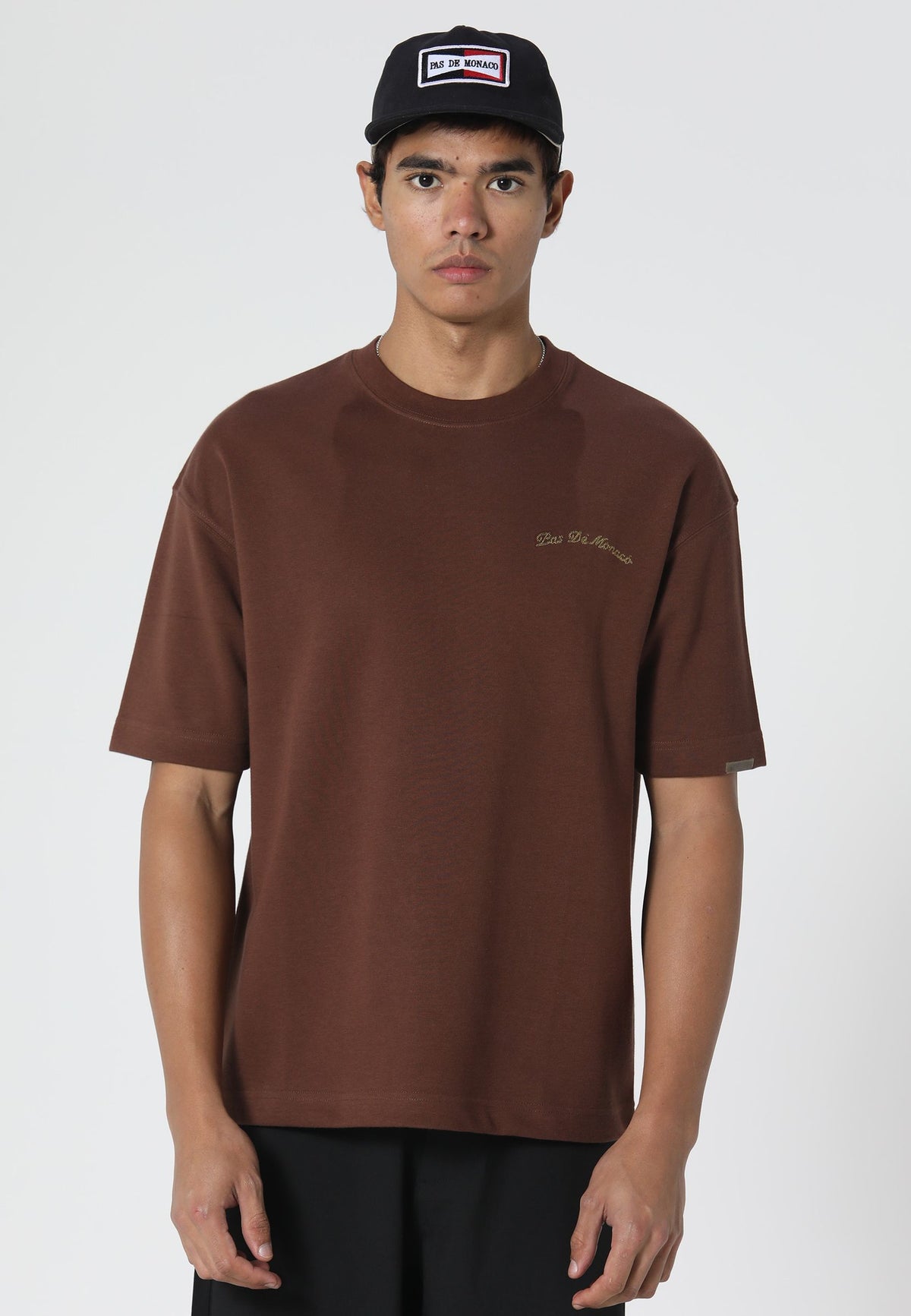 A man wearing a brown crewneck t-shirt with a small gold embroidered logo on the left chest, paired with a black baseball cap and black pants, photographed against a light gray studio background.