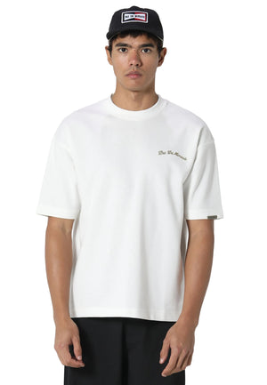 A man wearing a white short-sleeve crewneck t-shirt with a small gold embroidered logo on the left chest, paired with a black cap and black pants, photographed against a light gray studio background.