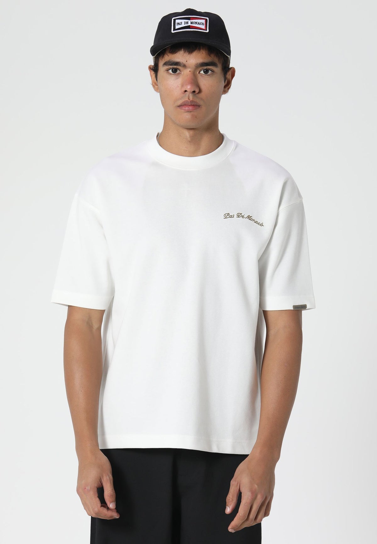 A man wearing a white short-sleeve crewneck t-shirt with a small gold embroidered logo on the left chest, paired with a black cap and black pants, photographed against a light gray studio background.