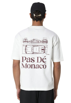 A man viewed from behind wearing a white t-shirt with a dark brown graphic of classic cars and the text 'Pas Dé Monacé' on the back, paired with a black baseball cap and black pants, against a light gray studio background.